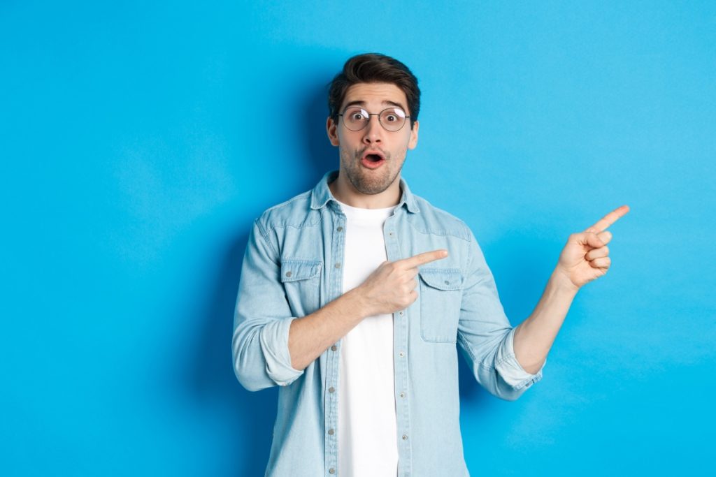 Homme surpris avec des lunettes pointant du doigt l'air intrigué, debout sur fond bleu.